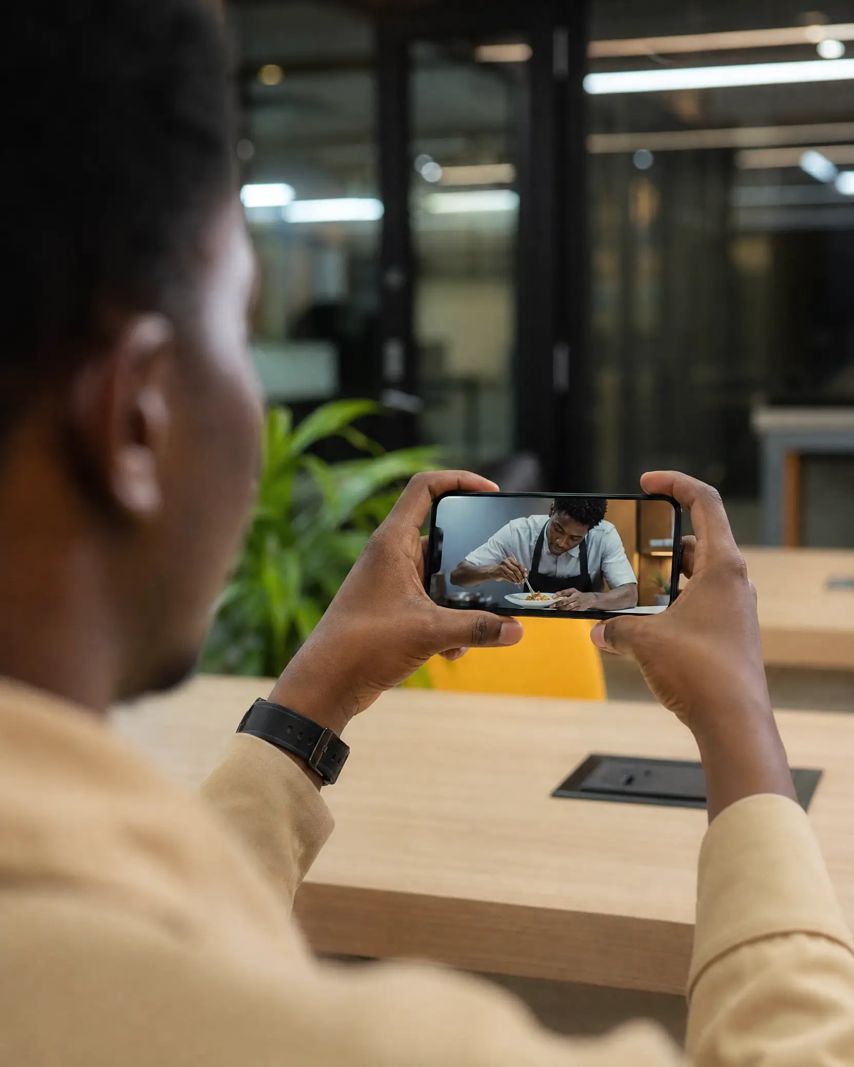 An over-the-shoulder view of a person in a beige shirt holding a smartphone horizontally, watching a video of a Black chef plating a dish at a wooden table.